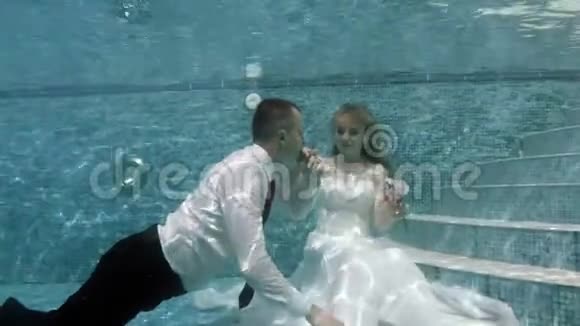 快乐的新郎和新娘穿着白色婚纱坐在池底的水下视频的预览图