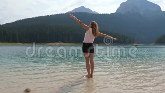 美丽的女人站在山湖的石头上双臂伸展到山坡上视频的预览图