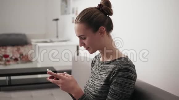 一位幸福的年轻女子坐在家里的沙发上看着电话打着一条短信的画像视频的预览图