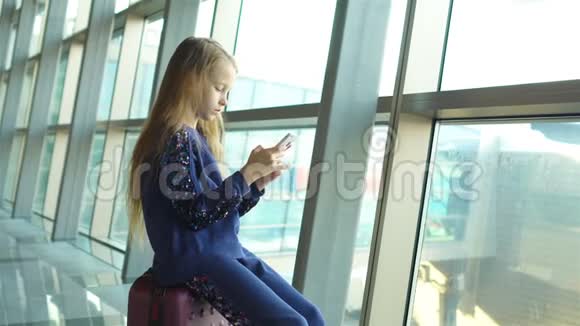 可爱的小女孩在机场附近的大窗户室内玩智能手机视频的预览图