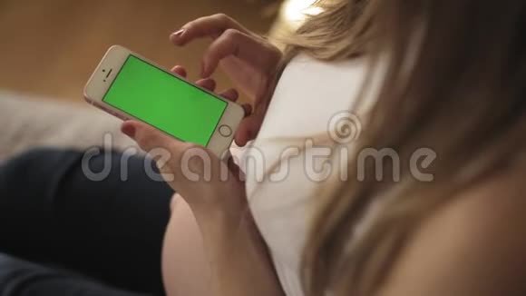 一位使用智能手机的女士的室内镜头绿色屏幕坐在白色沙发上滑动手势垂直方向视频的预览图