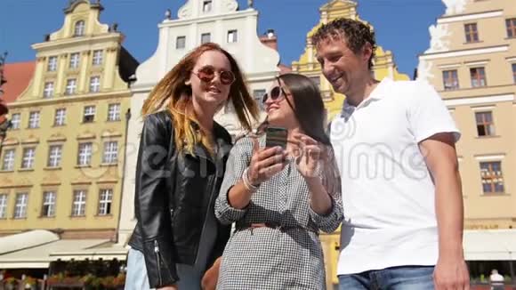 微笑的年轻人站在旧城广场上使用智能手机和触摸屏幕显示可爱的布鲁内特视频的预览图