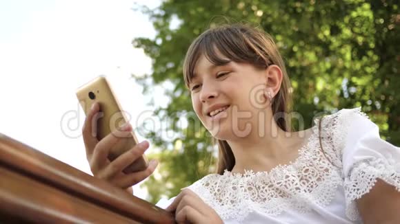 快乐女孩用智能手机在长凳上的公园里翻页特写镜头年轻的千禧年视频的预览图