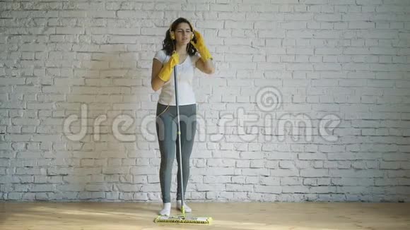戴着防护手套的漂亮年轻女子正在用拖把唱歌在客厅打扫房间时跳舞视频的预览图