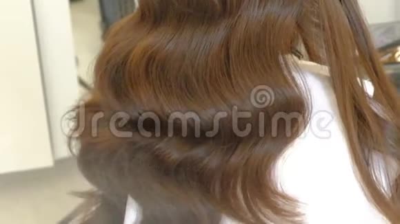女理发师带卷发铁的棕色长发在浅色美发沙龙的背面观看视频的预览图