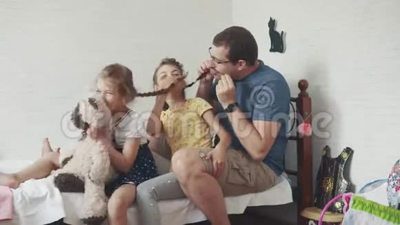 一位年轻的父亲和他的年幼的孩子玩耍爸爸和两个女儿坐在床上孩子们的房间里视频的预览图