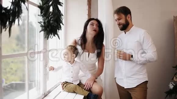 年轻可爱的一家人坐在装饰着圣诞花环的窗户旁爱的父母互相亲吻视频的预览图