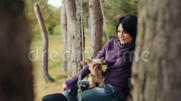 带着狗在长凳上荡秋千的女孩那个迷人的年轻女孩和狗坐在公园的长凳上慢动作视频的预览图