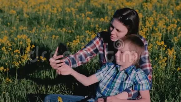 妈妈和儿子在手机上自拍坐在黄花间的草地上视频的预览图