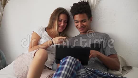 年轻的多民族夫妇在数码平板电脑上在床上笑着看电影慢镜头视频的预览图