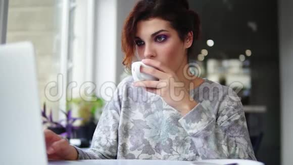 年轻漂亮的女人坐在靠近窗户的咖啡馆里喝咖啡用笔记本电脑工作女孩冲浪视频的预览图