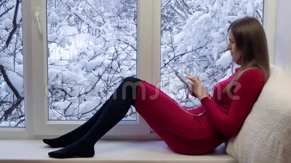 穿着红色裙子的漂亮女孩坐在窗台上看着窗户用带耳机的平板电脑冬天在外面视频的预览图