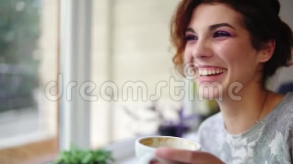 美丽的年轻女人在咖啡馆里坐在窗边享受咖啡和微笑女士配杯热气腾腾的饮料视频的预览图