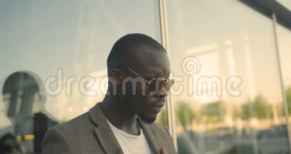 戴着现代太阳镜的时尚自信的非洲男人正在玻璃大楼附近修正他的夹克4K录像视频的预览图