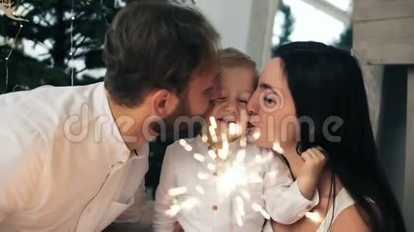 美丽的家庭新年快乐灯火辉煌年轻的父母亲吻他们可爱的孩子坐在旁边视频的预览图