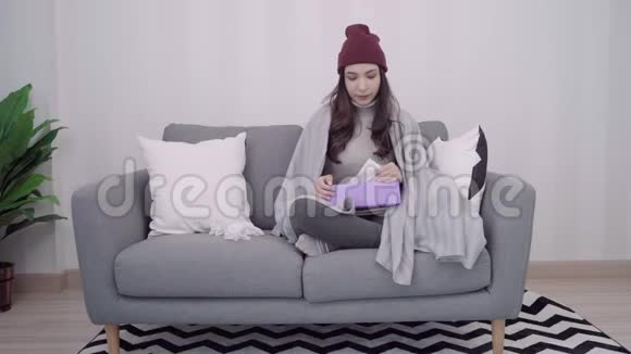 亚洲女人感到头痛裹着灰色毯子在客厅放松时一边躺在沙发上一边吹鼻子一边用纸巾视频的预览图