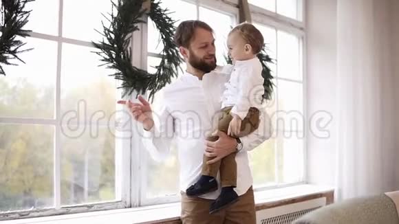 年轻的父亲抱着他的小男孩站在窗边看着外面窗户上装饰着圣诞节视频的预览图