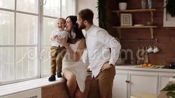 年轻的父母和他们可爱的儿子一起坐在窗台上拥抱和亲吻窗户是视频的预览图