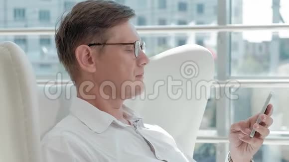 成熟的商人坐在玻璃办公室的白色椅子上用智能手机视频的预览图