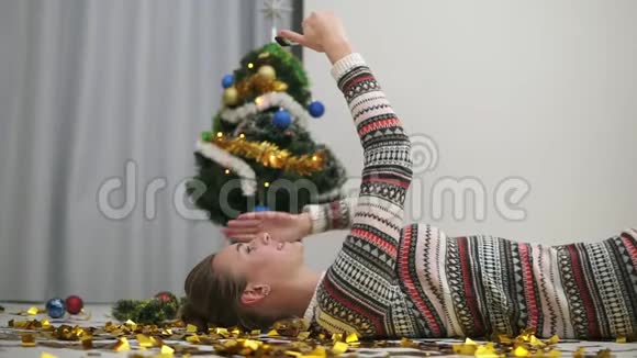 快乐的节日女孩躺在地板上的金色闪光纸屑和自拍发送吻装饰圣诞树视频的预览图
