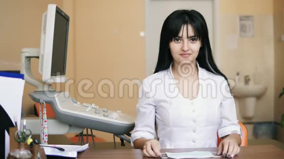 年轻微笑的女医生正在镜头前看着戴上一个保护性的外科口罩她坐在桌边视频的预览图