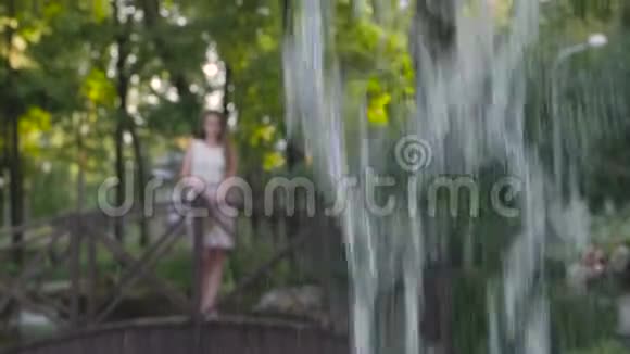 一个女孩走在桥上的公园里看着喷泉里涌出的水视频的预览图