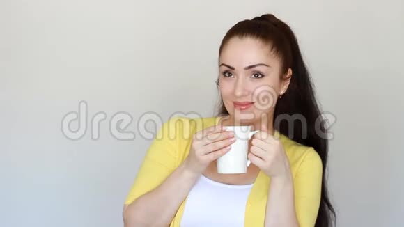 年轻的女人从杯子里喝一杯热饮料威奇握在他的手里女孩在演播室的白光背景上微笑视频的预览图