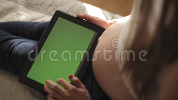 年轻孕妇坐在沙发上使用平板电脑和空白绿色屏幕视频的预览图