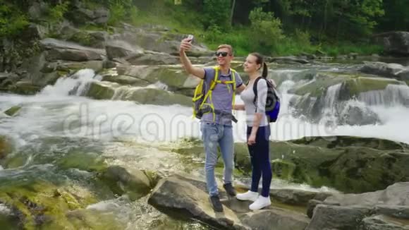 一对年轻的旅行者在山上瀑布的背景下自拍起重机抛丸视频的预览图