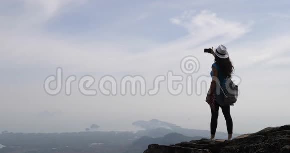 女游客背着背包站在悬崖上用手机拍摄山顶风景视频的预览图