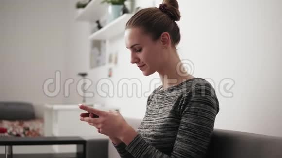美丽的年轻女子坐在家里的沙发上微笑着看着电话打字留言视频的预览图