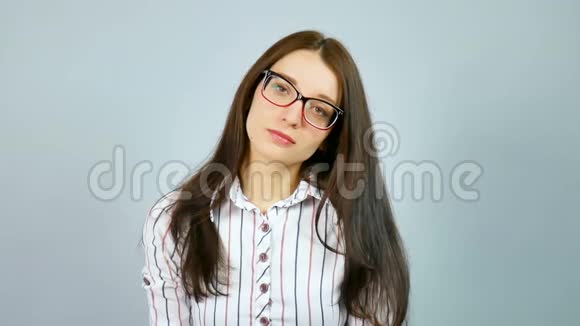 穿着红色和蓝色条纹的白色衬衫的严肃少女在灰色摄影棚看着她眼镜上的镜头视频的预览图