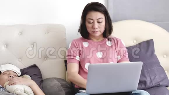 母亲带着一个孩子在家里试着用笔记本电脑工作一个生病的男孩躺在床上视频的预览图