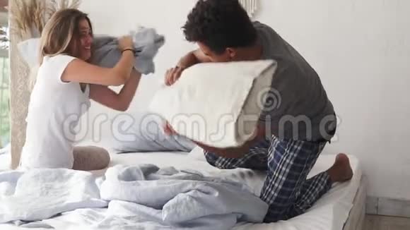 年轻的多民族夫妇在床上抱着枕头打架笑着摔倒真正的爱情观念早上好视频的预览图