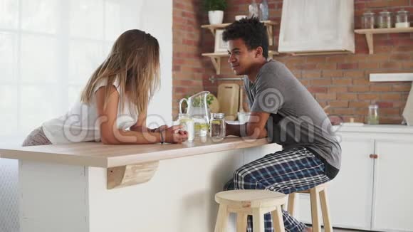 年轻的多民族夫妇早上在厨房的桌子旁交谈木桌上有水的玻璃视频的预览图