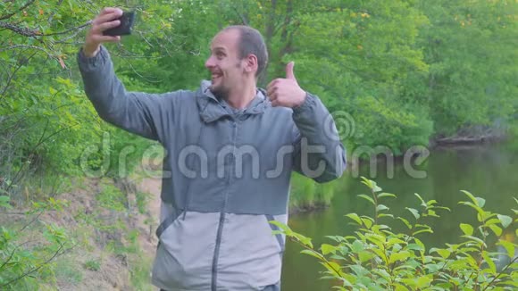 一个年轻人用河流和森林在风景中自拍看着镜头卷曲和鬼脸做一只手视频的预览图