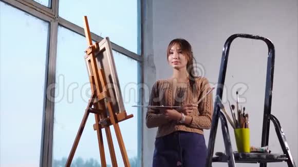 年轻的女艺术家站在画架附近拿着绘画工具站着视频的预览图