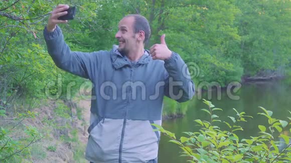一个年轻人用河流和森林在风景中自拍看着镜头卷曲和鬼脸做一只手视频的预览图
