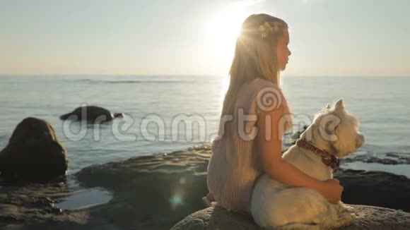 一个带着狗的悲伤的小女孩正坐在海边的石头上看着日落视频的预览图