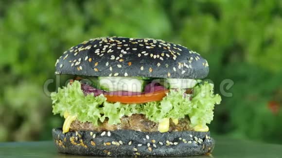 在绿色和蔬菜的模糊背景上带有黑色面包和芝麻的汉堡或汉堡包视频的预览图