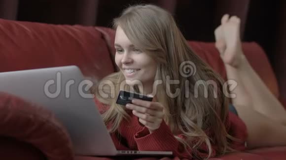 一位美丽的金发美女她靠在红色沙发上一边用笔记本电脑和信用卡购物视频的预览图