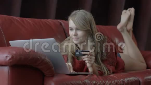 一位美丽的金发美女她靠在红色沙发上一边用笔记本电脑和信用卡购物视频的预览图