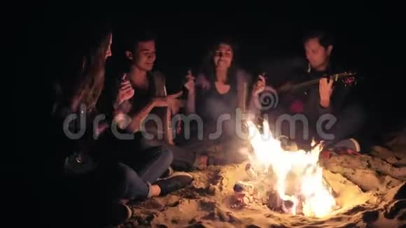 两个年轻的女人和两个男人深夜坐在篝火旁唱歌弹吉他和打击乐视频的预览图
