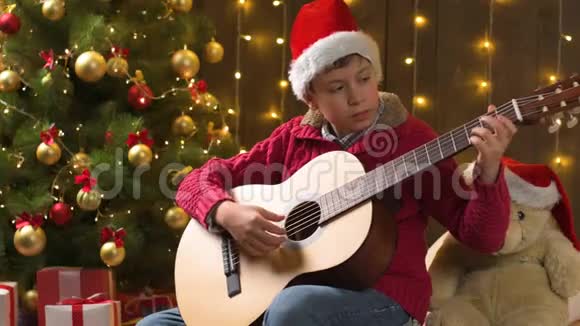 少年男孩弹吉他坐在室内靠近装饰好的圣诞树灯光打扮成圣诞老人助手圣诞快乐快乐视频的预览图