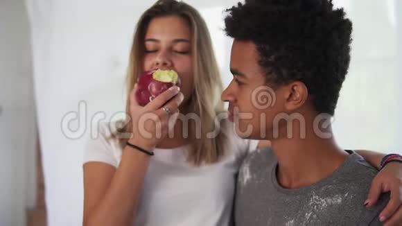 年轻的多民族夫妇在厨房做饭时互相拥抱美丽的白种人女孩吃苹果视频的预览图