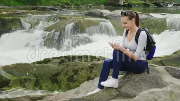 一个年轻的女人坐在山河背景下的岩石上使用智能手机技术和旅行概念视频的预览图