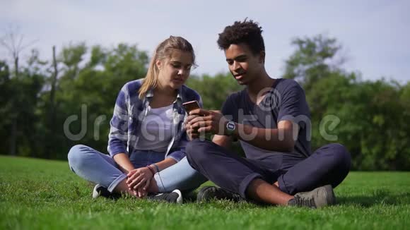混合种族夫妇坐在草地上在他们的智能手机中选择图片年轻人笑着笑可爱视频的预览图