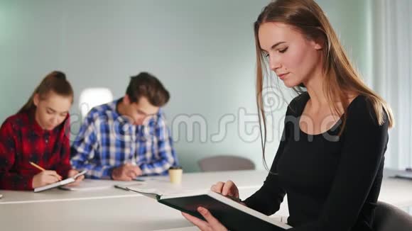 一位年轻的女学生在她的朋友正在学习时读一本书并做作业的画像视频的预览图
