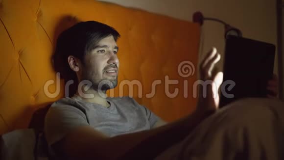 年轻的微笑男子使用平板电脑浏览社交媒体睡觉前躺在家里的床上视频的预览图