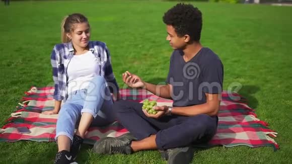 夏天一对年轻可爱的夫妇坐在公园的草地上吃水果男人小心翼翼地喂女人吃多汁的葡萄视频的预览图